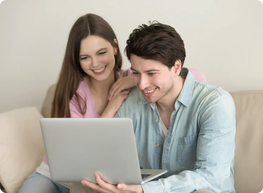 Young couple sitting together on a sofa, looking at a laptop screen together and smiling